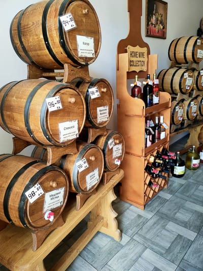 The image shows a retail display of wine and spirits in wooden barrels and bottles, likely within a shop or tasting room. The setting features rustic wooden barrels stacked on wooden stands, with some barrels equipped with spigots. Labels are visible on some of the barrels, indicating prices and potentially the type of beverage. Adjacent to the barrels is a wooden shelving unit displaying bottles of wine and spirits, with a rack at the bottom holding more bottles. A framed artwork depicting a still life is hung on the wall in the background. The floor is tiled with a wood-grain pattern. The overall aesthetic suggests a focus on artisanal or traditional beverages.