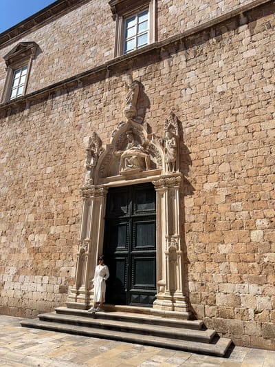 A person stands on the steps of a large stone building in Dubrovnik, Croatia. The building has a large, dark green double door with ornate carvings above it. There are also two windows visible on the upper part of the building. The person is wearing a white dress and white sneakers, and is looking at the camera. The ground in front of the building is paved with stone tiles. The overall scene is bathed in sunlight, casting shadows on the building and the ground.