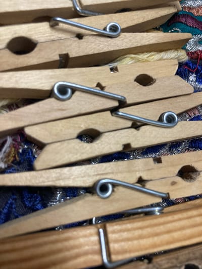 The image shows a close-up, overhead view of several wooden clothespins arranged diagonally on a patterned fabric. The clothespins are made of light-colored wood and have silver metal springs. Some of the clothespins are open, revealing the inner mechanism. The fabric beneath them has a complex embroidered pattern featuring blue, red, gold, and silver threads. There are no people or signs of Sabadell, Spain, visible in the image. The scene appears to be a still life composition.