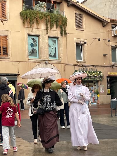 The image captures a group of people on a paved street in front of a building. One woman, wearing a large pink hat and a long white dress with pink trim, stands prominently. Another woman stands behind her in a long brown skirt and dark jacket with white swirls. Others are present, some holding umbrellas. The building behind them has windows with shutters and window boxes with flowers and plants. There's a sign on the building that reads "NATUR VELLA". Based on this sign and the architectural style of the building, it is likely in Terrassa, Spain, near the Natur Vella shop. The people in the image appear to be dressed in vintage clothing, possibly for a historical event or performance.