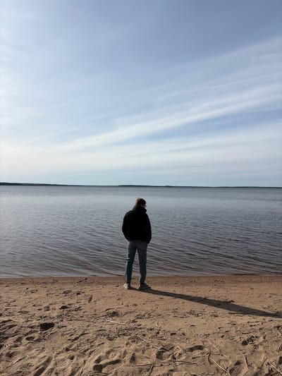 A medium-shot, vertically-oriented photograph shows a person standing on a sandy beach, looking out over a body of water. The beach takes up about one-third of the right side of the image, while the water occupies the central third, and the sky fills the left third. The person, facing away from the camera, is wearing a dark jacket and light-colored jeans. The sky is pale blue with streaks of white clouds. The water appears calm, with subtle ripples. The location is likely near the city of Glubokoe, Russia, given the context. The sand is tan and textured, with dry grass blades scattered around. A long shadow is cast by the person onto the sand.