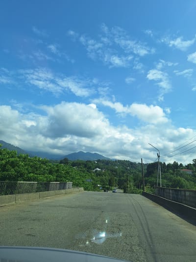 A road descends from a bridge, curving gently towards a distant mountainous landscape covered in lush green trees. The sky above is a vibrant blue, dotted with scattered white clouds, some appearing fluffy and others wispy. On the sides of the road, metal railings line the bridge, and further along, a few houses are visible nestled amongst the trees on the hillsides. A single car is seen on the road in the distance, and a lamppost stands on the right side, with power lines stretching across the frame. The road surface is asphalt and shows signs of wear, with cracks and patches evident. The overall impression is a scenic, somewhat rural setting.
