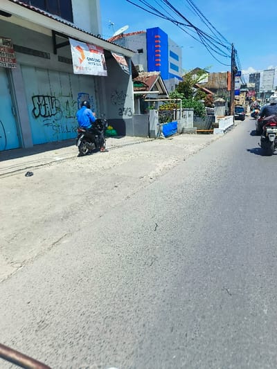 The scene is a street in Kecamatan Margahayu, Indonesia, with a single man riding a motorcycle on a paved road. The road appears to be relatively empty. The motorcycle is parked near the sidewalk on the right side of the road. The man is wearing a blue shirt and a helmet, and he is looking to the left. The road is wide and there is a small building on the left side of the road. The sidewalk is covered in gravel, and a small area is covered in grass. The building has a blue and white paint job and has a sign that says "GINO LOMO PIZZA" and a smaller sign that says "PROSPERA". There is a satellite dish on the roof. There are other houses, trees, and a power line in the background. The weather is sunny. The location is: -6.974 latitude, 107.573 longitude.