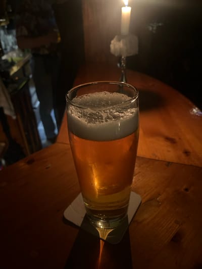 This media file presents a close-up, horizontally oriented view of a glass of beer resting on a wooden table, likely in a pub or bar setting in Bamberg, Germany. The beer, appearing light golden or amber with a substantial head of white foam, sits on a light-colored coaster. The glass reflects ambient light, highlighting the liquid's clarity.
To the left, a single lit candle in an ornate, possibly metal, candle holder casts a warm, soft glow across the scene, creating prominent shadows and illuminating the beer and table surface. The wooden table exhibits a rich, warm brown tone with visible grain, suggesting a rustic or traditional ambiance. The background beyond the table is dark and out of focus, implying a dimly lit environment, characteristic of an evening or nighttime setting.
The overall atmosphere conveyed is one of quiet relaxation and intimacy. No people are visible in the frame, and there is no discernible text. The focus remains on the drink and the subtle interplay of light and shadow provided by the candle.