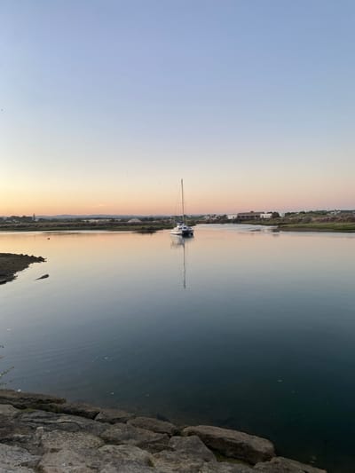 A sailboat is moored on a calm body of water in Olhão, Portugal. The sky above is a gradient of soft blues and oranges, suggesting either sunrise or sunset. The water reflects the sky and the sailboat, creating a mirror-like effect. In the background, a shoreline is visible with buildings, vegetation, and some piles of sand or construction material. The foreground features rough, dark rocks lining the edge of the water. There are no people or animals clearly visible in the scene.