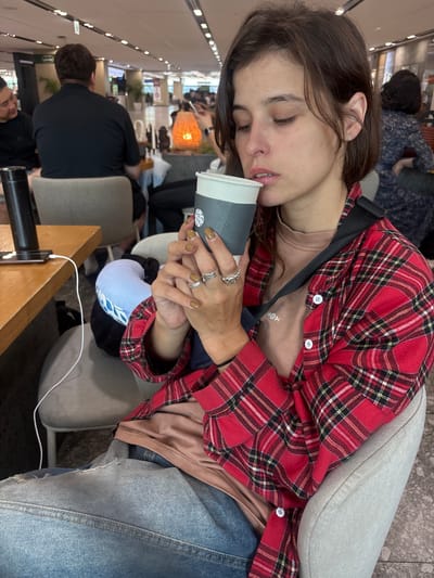 A woman with medium-length brown hair and closed eyes is sitting in an airport lounge, holding a coffee cup. She is wearing a red plaid shirt over a tan t-shirt and blue jeans, accessorized with rings on her fingers and a black strap across her chest. In the background, other travelers are seated and walking, with various seating areas and lounge amenities visible. The scene appears to be inside Jeju International Airport, given the presence of typical airport lounge features and other passengers.