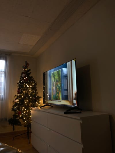 The scene depicts a dimly lit interior room in Bouxwiller, France, featuring a festive, domestic arrangement. A large, black, flat-screen television, exhibiting a yellow label on its upper right corner, rests centrally on a white, multi-drawer dresser. The television screen displays an image of two individuals, a male and a female, situated within a verdant, framed setting with architectural elements. A black television remote control is positioned on the dresser in front of the television. To the left of the dresser, a decorated artificial Christmas tree stands on a black base, illuminated by numerous warm-white string lights and adorned with red ornaments and other small decorations. On the far left, a window covered by sheer white curtains allows for minimal ambient light penetration. The room's ceiling features textured panels and a decorative molding strip along one edge. No individuals are present in the visible area of the image.