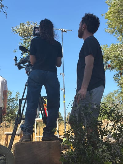 Two individuals are positioned outdoors on a bright, sunny day, likely in the Mate Yehuda Regional Council, Israel, given the vegetation which appears consistent with Mediterranean flora.
On the left, a person with long, dark hair, facing away from the camera, operates a professional video camera mounted on a tripod. They are wearing a dark t-shirt and blue jeans, standing on a wooden crate for height. The camera is equipped with various lenses and accessories, suggesting a filmmaking or photography session.
To the right, a person with curly dark hair and a beard, also facing away from the camera, stands near a metal pole, holding a bundle of leafy branches. This individual is wearing a dark t-shirt and light-colored shorts. The presence of branches may indicate a prop or set dressing element for the shoot.
The background features a clear blue sky and abundant greenery from trees and bushes, with some dry, yellowish vegetation in the mid-ground. The overall scene depicts a candid moment from what appears to be a film or photo production.