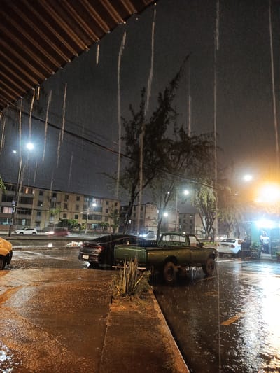 The photograph depicts a street scene during a heavy downpour at night, likely in Guayaquil, Ecuador. The foreground shows a wet sidewalk and street, reflecting the yellow-orange light from street lamps. Rain streaks are visible throughout the frame, indicating the intensity of the rainfall.

A green pickup truck is parked on the street alongside a dark-colored car. In the background, apartment buildings are visible. Streetlights illuminate the scene, casting a glow on the wet surfaces. A small business stall is positioned on the right side of the frame with at least one person. Other cars are also parked on the street. The overall atmosphere is dark and wet due to the heavy rain and nighttime setting.