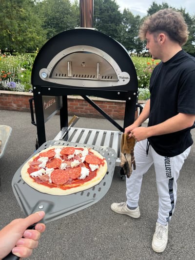An outdoor scene in Hornsea, United Kingdom, depicts two individuals engaged in pizza preparation on a paved surface. In the mid-ground, a black, dome-shaped "Vulcan+" outdoor pizza oven, featuring a stainless steel front with dual handles, a thermometer, and a chimney, stands on a black base with a lower metal shelf. To the right of the oven, a young male with light brown, curly hair, dressed in a black short-sleeved t-shirt, light grey sweatpants with "HOLLISTER" text on the left leg, and white athletic shoes, stands holding a pair of brown work gloves in his left hand while looking towards the oven. In the lower-left foreground, a partially visible hand holds a grey, perforated metal pizza peel, on which an uncooked pizza rests. The pizza features a red sauce base, topped with numerous slices of pepperoni and dollops of white cheese. The background includes a low brick wall, beyond which a vibrant garden with various green plants and colorful flowers is visible, backed by mature trees under an overcast sky.