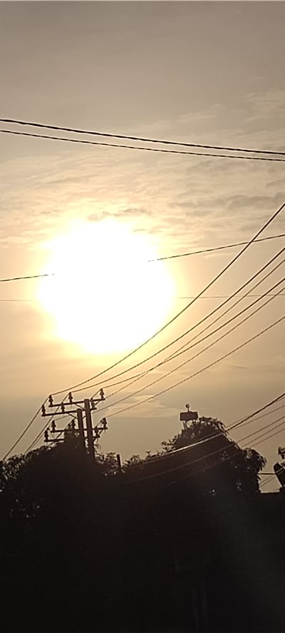 A wide-angle shot shows a bright sun in a cloudy sky, partially obscured by power lines. Below the power lines are trees and utility poles. The location appears to be Beunot, Indonesia. No people are visible.
