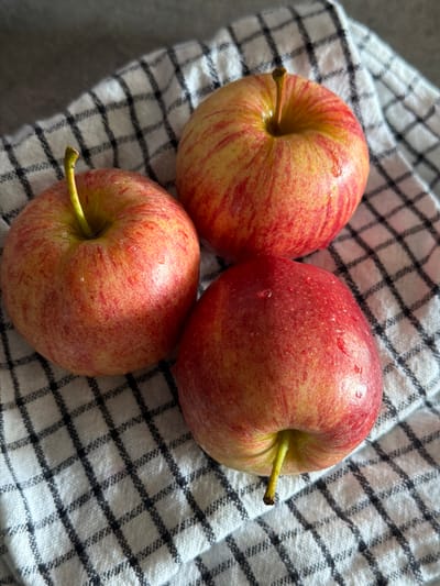 Three unpeeled apples, displaying red and yellow striped skin with scattered water droplets, are centrally arranged on a white textile featuring a black grid pattern. The apples are positioned with two in the upper frame and one below, forming a loose cluster. Each apple retains its green-brown stem. The underlying surface appears to be a grey, indistinct plane. The setting is an indoor environment in Champs-sur-Marne, France, characterized by soft, ambient illumination.