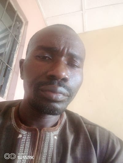 A close-up selfie of a dark-skinned man with a shaved head. He wears a brown, embroidered top. His expression is neutral. A window with metal bars is visible in the upper left corner. The wall behind him is painted a light peach color. The ceiling is white with rectangular patterns. The lower-left corner of the image features the "TECNO SPARK" logo. The man appears slightly sweaty.