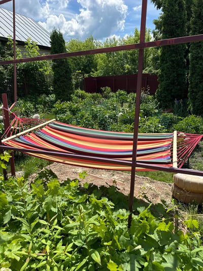 The image depicts a colorful striped hammock suspended between two metal posts in a lush green garden. The hammock is made of fabric with horizontal stripes in red, orange, yellow, green, blue, and purple. It is supported by wooden spreader bars at each end, and red ropes connect the spreader bars to the metal posts. In the background, there is dense foliage, including trees and bushes, with a red fence visible behind them. A portion of a building with a silver roof is also visible to the left. The sky above is blue with scattered white clouds. The scene appears to be a backyard or park setting, likely in a rural or suburban area. The location is stated to be Oryol, Russia, suggesting it is a summer day with warm weather conducive to outdoor relaxation.