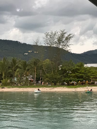 The image depicts a tropical beach scene in Nathon, Thailand. The foreground is dominated by the calm, green water of the ocean, reflecting the overcast sky. A sandy beach lines the edge of the water, dotted with small boats and vegetation. In the background, a lush, green mountain rises, partially obscured by clouds.
Several small boats are visible on the beach and in the water, including a green boat with an awning and a white inflatable boat. There are also what appear to be temporary structures or stalls set up along the beach, with yellow and white awnings. Palm trees and other tropical foliage are scattered across the beach and near the base of the mountain, suggesting a warm, humid climate.
The sky is heavily clouded, indicating either an overcast day or potentially approaching rain. The lighting is soft and diffused, consistent with a cloudy day. The time of day is difficult to ascertain precisely due to the lack of direct sunlight and shadows, but it appears to be daytime.
There are no visible people actively engaged in a specific activity, though some figures may be present near the structures on the beach. The overall mood of the image is serene and tranquil, with the natural beauty of the tropical landscape being the primary focus. The image is taken from a slightly elevated perspective, looking down towards the beach and the water. There is no visible text in the image.