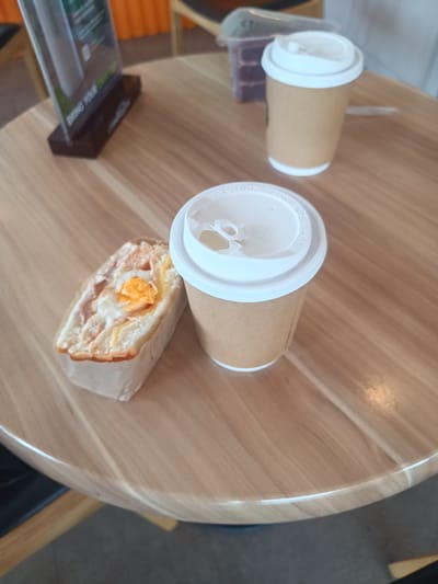 On a round wooden table in an indoor setting, likely a cafe or restaurant, two paper cups with white lids are present. Next to one of the cups is a sandwich in a paper wrapper, revealing an egg and sausage filling. In the background, a menu stand is visible, along with what appears to be a container of cake. The scene is in Plai Phraya, Thailand. No people are visible. The subjects include the food and drinks. The objects are the cups, the sandwich, the table, the menu stand and the container of cake. There are no actions or interactions occurring in the image. The environment is a cafe or restaurant setting with wooden furniture.