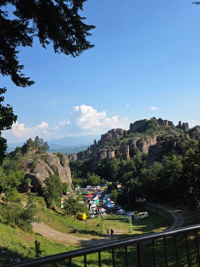 The image depicts a scenic outdoor gathering at a rocky, forested location, likely a tourist area or campsite. The scene is dominated by rugged, weathered rock formations covered in lush green trees, creating a dramatic natural backdrop. Below the rock formations, a grassy area hosts a collection of tents and a few vehicles, suggesting a camping event or festival. Several colorful tents and awnings are set up, with some people visible walking among them. A bright yellow portable unit is positioned on the grass, and a green metal structure, possibly a stage or shelter, is also present. A winding path leads through the area, and a railing, suggesting a viewpoint or walkway, is visible in the foreground. The sky is a vibrant blue with scattered white clouds, indicating fair weather during daytime. The overall impression is one of a lively event set within a beautiful and imposing natural landscape.