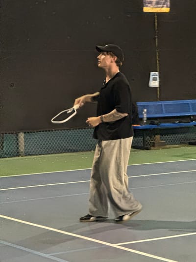 The image depicts a person standing on a sports court, likely for tennis or pickleball, at night. The subject is an adult male, captured in a full-length shot. He wears a black baseball cap, a black short-sleeved t-shirt, and wide-leg light grey pants with vertical ribbing or stripes. Tattoos are visible on his left arm. He holds a white-framed paddle or short-handled racket in his right hand. His head is turned to the left, suggesting he is looking forward or observing something off-frame, with a focused or neutral expression.
The court features distinct sections of blue and green with white boundary lines. In the background, a dark wall on the left side of the frame displays two pieces of paper or signs, though their text is illegible. To the right of the person, a chain-link fence separates the playing area from a blue metal structure, which could be part of bleachers or a facility support. A water bottle is visible resting on this blue structure.
The scene is illuminated by artificial light, indicating nighttime. The court surface and the fence show some reflective spots, possibly due to dampness or specific lighting conditions. The person's attire and the presence of the paddle suggest an activity related to racket sports. The overall setting is consistent with a recreational sports facility, located in West Hollywood, United States of America.