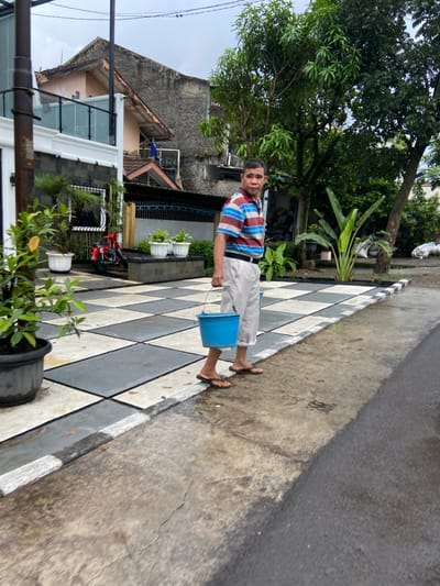 A man in a striped shirt and khaki pants is walking on a checkered concrete path towards the camera. He carries a blue bucket in his right hand. There is a motorcycle parked in the background. The path leads to a street with a black asphalt surface, the street appears to be wet. The surrounding environment is a residential area in Margaasih, Indonesia, with houses, trees, and plants. The latitude and longitude of the scene are -6.944 and 107.555, respectively.