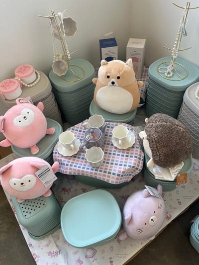 This image displays a retail merchandise arrangement in Edmond, United States. The scene is presented against a plain white wall, with merchandise resting on a light-colored floor partially covered by a white, illustrated runner.
The central focus is a miniature tea set positioned on a checkered fabric (pink, blue, white, gray) laid over a light teal, ribbed storage bin. The tea set includes a glossy, iridescent purple teapot and four white, scalloped teacups with matching saucers.
Surrounding this central display are various plush toys and storage containers. To the left, two round pink plush toys, resembling pig-like figures with white faces, are stacked on light teal and gray ribbed storage bins; the lower pink plush bears a product tag. A light brown, dog-like plush toy is centrally located in the upper mid-ground, seated on a stack of light teal bins. To the right, a brown hedgehog-like plush toy is positioned on additional light teal bins, also displaying a product tag. In the lower right foreground, a light purple, round plush toy resembling a cat or similar animal rests on the floor.
Two gold-colored metal jewelry stands are visible, one on the left and one on the right, each draped with multiple strands of white pearl necklaces and holding small, white scalloped dishes. Further to the left, two small, pink circular containers with embossed floral lids are positioned on stacked gray bins. Several white cardboard product boxes, some with discernible "Disney" branding, are visible in the background behind the dog-like plush toy. The storage bins are consistently light teal or gray, featuring vertical ribbing, and are stacked to create varying display heights.