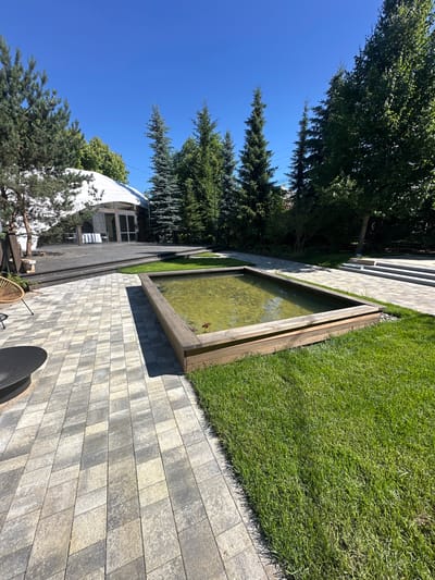 A rectangular, raised pond with wooden borders is situated in a paved outdoor area. The pond's water is murky green, and the bottom is visible, with what appear to be small stones or pebbles. Adjacent to the pond is a meticulously manicured lawn. In the background, a large, white, tent-like structure with glass doors and windows stands on a dark wooden deck. Several evergreen trees, including tall, slender ones and fuller pines, surround the area. The sky is a clear, bright blue, indicating a sunny day. The scene is likely an outdoor event space or a well-kept garden in Zakharovo, Russia.