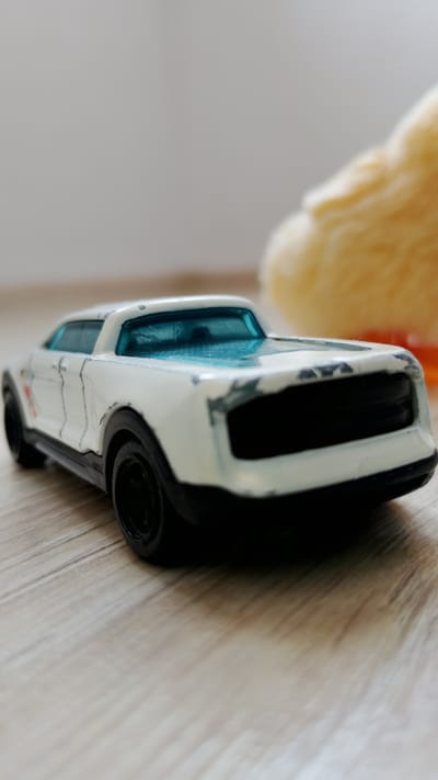 This is a close-up, eye-level shot of a white toy car with a blue windshield, placed on a light-colored wooden surface. The car is angled towards the right, with its front facing the viewer. The toy car appears to be a model of a futuristic or retro-futuristic automobile, possibly a modification of a classic muscle car, with a wide grille and sharp, angular lines. The paint on the car shows some signs of wear and tear, with scuffs and chips visible on the edges and hood. To the right of the car, out of focus, is a fuzzy, yellow object, possibly a plush toy. The background is a plain, light-colored wall. The lighting is soft, casting gentle shadows on the wooden surface. There are no people or other discernible objects in the frame, apart from the toy car and the out-of-focus yellow item. The image is taken indoors, likely in a residential setting. There is no information or visual evidence to suggest the location is in the city of Smolyan, Bulgaria.