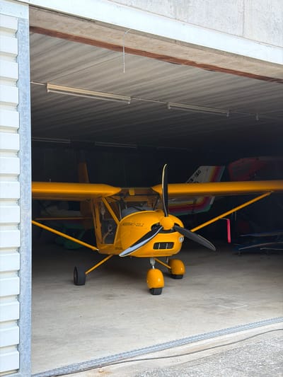 The image depicts the interior of an aircraft hangar, likely located in Becherbach, Germany. A bright yellow, single-engine ultralight aircraft, identified by the text "AEROPRAKT-22L2" and "ULTRALEICHT" on its fuselage, is the primary subject. The aircraft is positioned centrally within the hangar, facing forward. Its propeller is visible, along with its front landing gear, which consists of two small wheels and fairings, and a single tailwheel. The hangar itself is constructed with corrugated metal siding on the exterior and a corrugated metal roof. Fluorescent lighting fixtures are mounted on the ceiling. In the background, partially obscured by the yellow aircraft, are other aircraft or aircraft parts, including a tailfin with a rainbow-colored stripe and some red and blue elements. The ground is a concrete floor, with some white markings. The hangar door is partially open on the left side, revealing a glimpse of the exterior. There are no people visible in the image.