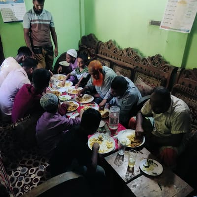 A group of approximately ten to twelve males, spanning various ages from children to older adults, are gathered indoors for a communal meal. The setting appears to be a home or local establishment in Lalmohan, Bangladesh, with light green walls and ornate wooden couches. The time of day seems to be evening or night, indicated by the primary reliance on artificial lighting.

Most of the individuals are seated on the floor or low cushions around a low, rectangular wooden table, actively eating from white plates using their hands. The plates contain rice and what appears to be a meat or curry dish. Glasses of water and a large pitcher are distributed on the table, alongside a plate of sliced cucumbers and green chilies.

Notable subjects include an older man with a distinctive orange-red beard, several younger men in casual attire, and children, one of whom wears a white taqiyah (skullcap). In the background, a man stands, observing the meal, dressed in a polo shirt and a patterned lungi.

On the left wall, a calendar with Bengali script is visible. Another document or calendar, also with Bengali text and numbers such as "২" (2) and "৩১" (31), hangs on the right wall above an electrical switchboard. The floor is covered with a dark patterned rug. The scene conveys a relaxed and intimate social gathering around a shared meal.
