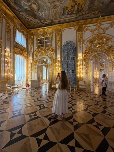 This image depicts the interior of the Catherine Palace, located in Pushkin, Russia. The scene is a grand, ornate hall characterized by lavish gold gilded decorations, high ceilings with painted frescoes, and a polished wooden parquet floor with a geometric pattern of light and dark wood. A large, blue and white tiled stove or fireplace stands prominently against one wall.
In the foreground, a woman with long, brown hair is standing with her back to the camera, holding a smartphone and appearing to take a photograph. She is wearing a long, flowing white skirt with a blue trim at the hem and light brown shoes.
Further into the hall, another woman is visible on the right side of the image, also looking towards the interior of the palace. She is wearing a white t-shirt and dark pants, and is carrying a light-colored bag over her shoulder.
Several elegant chairs upholstered in white are placed around the hall, and brass stanchions with ropes are set up to guide visitors. Ornate candelabra with multiple light sources are mounted on the walls, contributing to the rich and illuminated atmosphere of the room. The overall impression is one of opulence and historical grandeur.