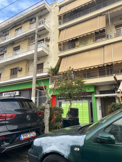 This image captures a street scene in Koridallos, Greece, during what appears to be daytime, under a partly cloudy sky. The photograph, presented with a counter-clockwise rotation, shows a mix of residential and commercial architecture, parked vehicles, and natural elements.
On the left side of the scene, a multi-story residential building dominates, featuring a light-colored facade with some signs of wear and peeling paint. Numerous balconies with white railings are visible, many of which have extended beige fabric awnings, suggesting protection from the sun. Several air conditioning units are mounted on the building's exterior.
Below the residential structure, a commercial establishment occupies the ground floor, distinguished by a bright green facade with a horizontal red band near its base. A large window displays prominent text, "BOX NOW," indicating a delivery or locker service, along with other graphical elements.
In the foreground, two cars are parked parallel to the curb. On the right, a dark green car is partially visible, showing its front fender and a portion of its windshield and side mirror. Next to it, a black MG SUV or hatchback is parked, its rear visible. The vehicle's license plate displays "XHA-9742" with a Greek flag symbol, confirming the location. The word "entopizw" (Greek for "I locate" or "I track") is also visible on the rear of the black car.
Several trees and shrubs are planted along the sidewalk, providing greenery to the urban setting. A notable tree with a distinct, gnarled trunk stands between the commercial building and the parked cars. No people are visible in the scene, which appears to be quiet and still.