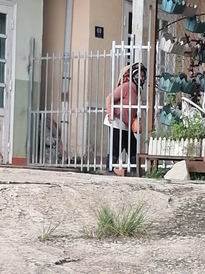 A person wearing a pink hoodie and black pants is standing behind a metal gate in Xuân Thành, Vietnam. The person is wearing a headscarf and a mask. In the background, there is a house with a number 69 on the wall and hanging planters. The ground is paved and there is some dry grass growing in cracks.