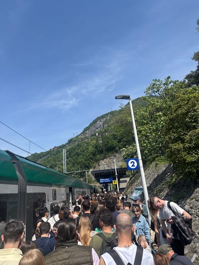 The image depicts a crowded train platform at the Varenna-Esino-Perledo train station in Gittana, Italy. Passengers are densely packed near the train doors, presumably boarding or disembarking. A green and silver train is visible alongside the platform. A sign indicates the platform number '2'. Beyond the platform, a steep hillside with vegetation and some visible structures rises into the background. The sky is clear and blue. A light pole stands near the platform edge, and a stone wall runs along the upper part of the scene. Several people are seen standing or sitting near the wall.