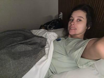 The image depicts a woman lying in bed, facing the camera, with a portion of her body covered by a grey blanket and white and light green sheets. She has dark hair, light-colored eyes, and is smiling. Her left arm is extended, and her right shoulder and upper arm are visible. In the background, a dark wooden headboard is present, along with electrical outlets and some charging cables, suggesting the image was taken in a hotel room in Incheon, South Korea.