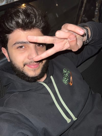 A young man with dark curly hair and a beard is posing for the camera. He's wearing a black hoodie with a Nike logo in green and orange. He is flashing the peace sign. He has a silver bracelet on his wrist. The background appears to be the interior of a car.