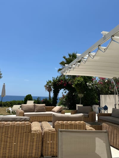 This is a wide outdoor shot taken from a ground-level perspective, showcasing a patio area with rattan furniture and a view of the sea. In the foreground, a collection of plush, tan-cushioned rattan sofas and armchairs are arranged, some with woven detailing. Behind the furniture, a lush green lawn leads to a white retractable awning supported by a metal frame. Beyond the lawn, the deep blue Mediterranean Sea is visible under a clear, bright blue sky. A few palm trees dot the landscape, and a white patio umbrella stands closed on the left. A stone archway covered in vibrant pink and white flowers frames the view of the sea and foliage. The scene appears to be a relaxation or dining area at a resort or private residence in the Kissonerga Community, Cyprus, given the typical climate and vegetation suggested by the image. There are no people or animals visible in the frame.