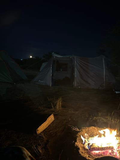 A nighttime scene depicts a campsite with two tents and a campfire. The larger tent, a modular camping shelter with a grey fabric exterior, is situated in the center of the frame. A smaller, green tent is positioned to the left. The campfire, burning brightly with orange and yellow flames, is in the foreground on the right. Dry grass and sparse vegetation surround the tents and campfire on a dirt surface. The sky above is dark, with a faint moon visible in the distance. The location is identified as Колодезный, Russia.