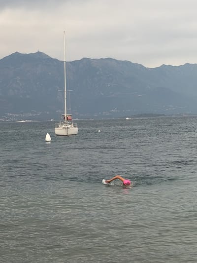 In this vertical, outdoor, daytime photograph, a person is swimming in a body of water with mountains in the background. The swimmer, wearing a pink swim cap and a pink swimsuit, is in the right-center of the frame, with their left arm extended in the water, creating a splash. The water is a dark, choppy grey, reflecting the overcast sky. In the left-center of the frame, a white sailboat with a tall mast is moored. The sailboat is white and appears to have some red accents on its deck. A white buoy floats near the sailboat. The mountains behind the water are dark and steep, covered in a muted green landscape, and rise towards a pale, cloudy sky. The scene suggests a calm but slightly cool day. The location appears to be a coastal or lakeside area, with the mountains indicating a mountainous region.