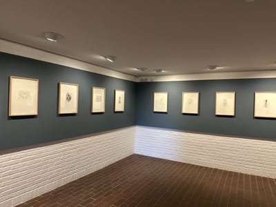 The scene depicts an interior view of an art gallery or museum space, identified as being in Humlebæk, Denmark. The room features a white ceiling containing multiple recessed circular downlights. The walls are horizontally divided: the upper section is painted a dark teal color, while the lower section, approximately 1.2 meters in height, consists of white-painted brickwork. A thin white trim separates the painted wall from the brickwork, and a wider white trim borders the ceiling. The floor is composed of reddish-brown square tiles arranged in a grid pattern.
Eight framed artworks are displayed. Four vertical rectangular frames, constructed of light-colored wood with white matting and containing monochromatic drawings or prints, are evenly spaced on the left wall. The leftmost artwork depicts two stylized hands or a symmetrical open form. The second shows an abstract organic shape. The third contains a faint geometric or textural pattern. The fourth portrays a singular, stylized object resembling a chess piece or vessel. Four additional similarly framed artworks are visible on the perpendicular wall to the right. No individuals are present, and no actions are occurring within the depicted environment.