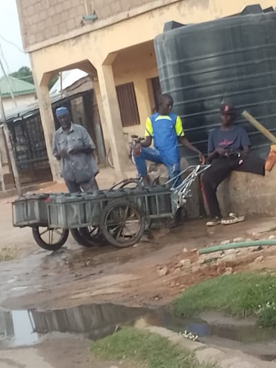 In Arawa, Nigeria, three individuals are gathered near a large black water tank. On the left, an older man in a blue cap and grey shirt stands beside a cart filled with what appear to be batteries. In the center, a younger person wearing a blue and yellow top and jeans is seated on the cart's frame. To the right, another young person in a dark t-shirt and black pants is seated on a concrete ledge. The scene takes place outdoors, with a building in the background and a muddy area with a puddle in the foreground.