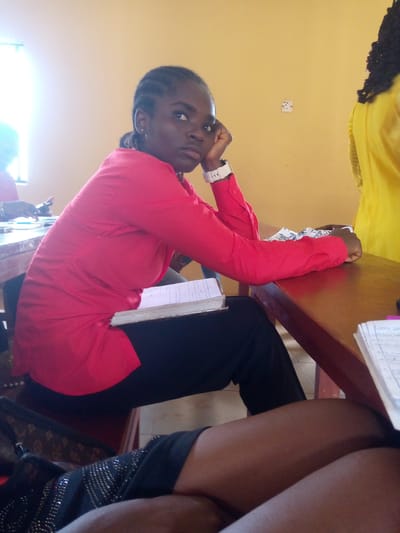 This is a medium shot of a young Black woman with braided hair, wearing a bright red collared shirt and black pants, sitting at a wooden desk in what appears to be a classroom setting in Nsugbe, Nigeria. She is looking to her right with her chin resting on her hand, appearing thoughtful or bored. A white wristwatch is visible on her left wrist. A book lies open on the desk in front of her. Another person, mostly out of focus and wearing a bright yellow garment, is visible standing behind her and to the right. To the left, another individual's hand is visible, resting on the desk. A patterned cloth is also on the desk. The wall behind the subjects is a pale yellow, and a power outlet is visible on it. The lighting suggests an indoor environment, possibly illuminated by natural light from a window on the left side of the frame. The overall scene suggests a moment of study or a break during a class.