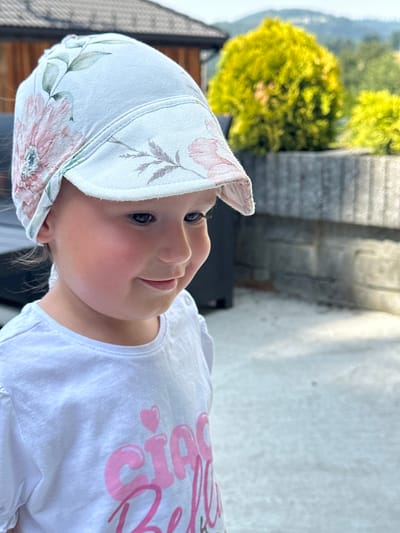 A young child is captured in a close-up, outdoor portrait, with the image rotated 90 degrees clockwise. The child, wearing a white cap with a floral pattern of pink and green flowers and leaves, looks to the right of the frame with a gentle smile. They are also wearing a white t-shirt with large, puffy pink lettering that appears to spell out "GIRL" with a small heart next to the "G". The background is softly blurred, showing hints of greenery from trees and a stone wall, suggesting a bright, sunny day in a natural or garden setting. The lighting suggests it is daytime, possibly late afternoon given the warm glow. The child's expression conveys a sense of joy and contentment. The setting appears to be outdoors in a pleasant, possibly residential or park-like area, with elements that could be associated with Wisła, Poland.