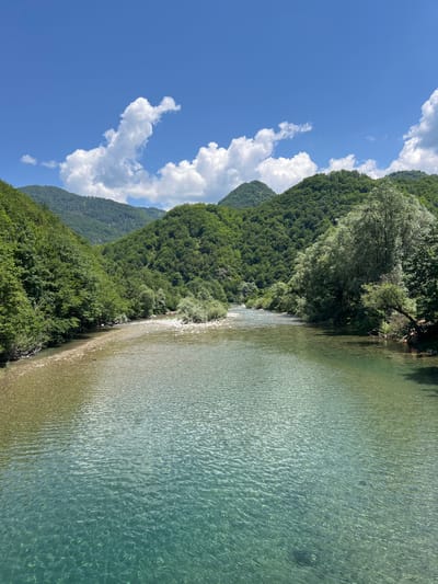 This is a photograph of a clear, shallow river flowing through a valley in Moračko Trebaljevo, Montenegro. The riverbed is visible through the crystal-clear water, revealing a gravelly bottom. Lush green trees and vegetation cover the rolling hills that flank the river. The sky above is a vibrant blue, dotted with fluffy white clouds. The scene is one of natural beauty and tranquility.