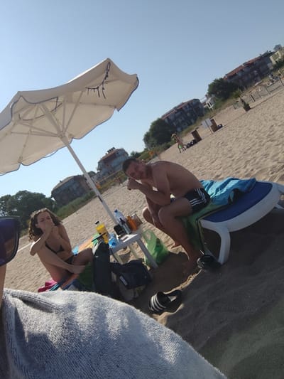 The image depicts a sunny day at a beach in Lozenets, Bulgaria. In the foreground, a person, whose face is not fully visible, is covered by a gray towel. To the left, a woman with curly brown hair, wearing a black bikini, is sitting on the sand, holding her hand near her mouth. Beside her, a white beach umbrella provides shade. In the middle ground, a man with short brown hair, wearing black swim trunks, is sitting on a blue and white lounge chair, with a blue towel draped over it. A small white table is placed in front of him, holding a yellow bottle, a blue water bottle, and other items. In the background, several buildings and trees are visible, along with a volleyball net. The sandy beach extends to the horizon under a clear blue sky.