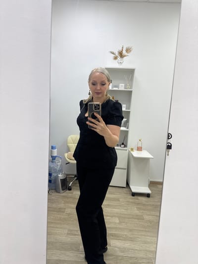 A woman with blonde hair styled in a ponytail takes a selfie in a mirror. She is wearing a black top and black pants. The background shows a room with shelves, a chair, and a small table with decorative items. There is also a water cooler and a small heater visible. The floor is made of light-colored wood. The woman is holding a smartphone in her right hand, which is capturing her reflection. The scene appears to be in an indoor setting, possibly a salon or office. The location is Komsomolsk-on-Amur, Russia.
