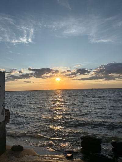 The image shows a wide expanse of choppy, dark blue water under a bright, partly cloudy sky. The sun, a brilliant orange disc, is setting on the horizon, casting a shimmering golden path across the water's surface. Wispy white clouds streak across the upper sky, while darker, heavier clouds are clustered around the setting sun. In the foreground, a concrete structure with weathered white paint and exposed brickwork juts out from the left side of the frame, partially obscuring the shoreline. The shoreline itself is sandy and wet, with a few dark rocks and discarded car tires visible. The overall scene captures a tranquil yet dramatic sunset over the sea, likely near a coastal area.