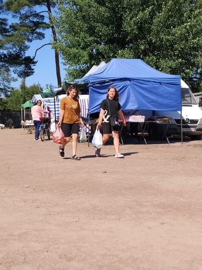 Two women are walking in a market setting. The woman in the foreground on the right is wearing a black t-shirt and black shorts, carrying a white plastic bag with items. She has a black crossbody bag. The woman on the left is wearing a brown t-shirt and black shorts, carrying a pink plastic bag filled with produce. In the background, a woman in a pink shirt is standing near a bicycle and a market stall. There are blue and green market tents, tables with goods, and a white van. The ground is dirt. The setting appears to be an outdoor market. The license plate on the van reads "AM 8339 AO". Based on the license plate format, this vehicle is likely registered in Ukraine. Irshansk is a city in the Zhytomyr Oblast of Ukraine, consistent with the presence of Ukrainian vehicles.