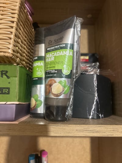 A shelf in Perushtitsa, Bulgaria, holds several hair care products. In the foreground, a clear plastic-wrapped tube of "Dr. Santé Macadamia Hair Conditioner" is prominent, featuring images of macadamia nuts and green leaves. To its left is another, similar hair product tube, partially obscured by the first. Behind these, a woven basket is visible on the upper left. To the right of the hair products, a black, rough-textured object with torn edges sits on the shelf. The shelf itself is made of light-colored wood and is part of a larger wooden structure, suggesting it's in a room with wooden furniture. The lighting is dim, creating shadows.