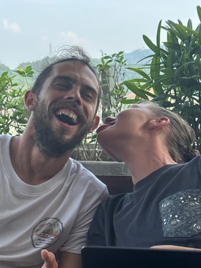 The image captures a lively outdoor moment featuring two individuals in what appears to be Hà Giang 1 Ward, Vietnam. In the foreground, a man with a dark beard and curly hair is shown mid-laugh, his mouth wide open, displaying his teeth and an expression of pure joy. He is wearing a white t-shirt. From the upper right side of the frame, another person, whose hair is pulled back, leans playfully towards the laughing man, sticking out their tongue. This person is dressed in a dark grey t-shirt that features a pixelated camouflage-like pattern on its left breast pocket.
The background is filled with lush green foliage, including large palm-like leaves, suggesting a tropical or mountainous environment. Beyond the immediate greenery, the distant landscape is hazy and light, hinting at misty mountains or hills under an overcast sky, consistent with the specified Vietnamese location.
On the man's white t-shirt, a circular graphic design is visible. Text within this graphic clearly reads "HA GIANG" and "VIETNAM". Other discernible elements include numbers like "346.3" and "FT" or "FEET", likely indicating an elevation or specific location marker, along with the word "AND". The overall scene evokes a sense of lightheartedness and friendly interaction amidst a beautiful natural backdrop. The time of day appears to be daytime, possibly overcast or hazy.