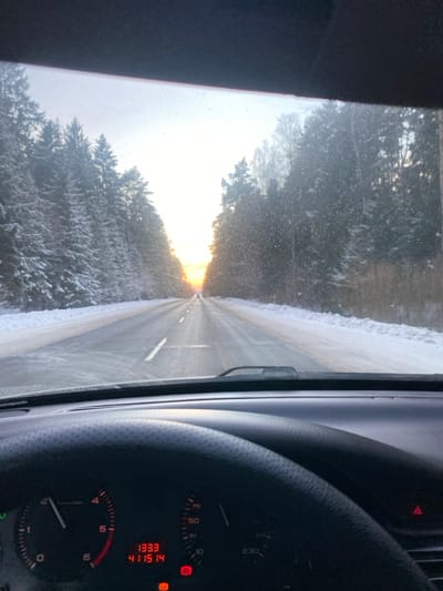 The image presents a view from inside a vehicle on a winter road in Карабоўскі сельскі Савет, Belarus. The perspective is forward through a windshield partially obscured by small particulate matter, possibly snow or ice. A two-lane asphalt road, cleared of snow in its main thoroughfare but with snow-covered shoulders, extends into the distance. It is bordered on both sides by dense conifer forests laden with snow.
A low sun, characterized by an intense orange glow, is visible directly ahead on the horizon, casting strong light and a slight lens flare. The sky above is bright and hazy.
In the foreground, the vehicle's dashboard is visible. A portion of a black, textured steering wheel is on the lower-left. Behind it, the instrument cluster displays a tachometer needle at approximately 1000 RPM and a speedometer needle at 0 km/h. A digital readout shows "13:33" and "411514". Several red warning lights are illuminated on the dashboard, including a circular symbol with an exclamation mark. Faint lights from distant vehicles are discernible on the road ahead, partially obscured by the sun's glare.