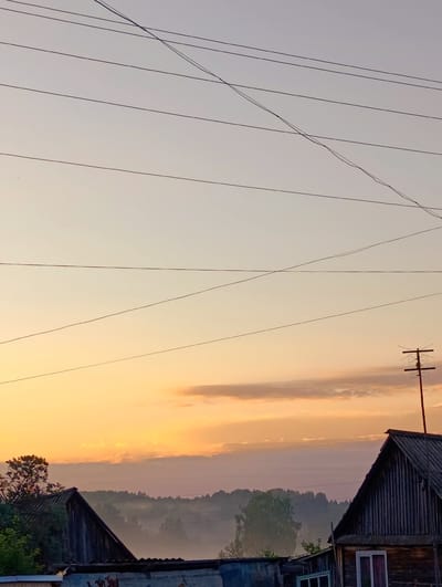 The image depicts a rural scene at sunrise, with a soft gradient of orange and pale yellow in the sky. Several parallel power lines stretch across the upper portion of the frame. In the foreground, the roofs of two rustic wooden houses are visible. The house on the right has a dark, corrugated roof and a visible window with pinkish curtains. A TV antenna on a red pole stands atop this house. The house on the left, partially obscured, appears to be made of weathered wood planks. A line of trees and rolling hills is visible in the mid-ground, partially shrouded in a light mist. The overall impression is one of a quiet morning in a village setting, likely in a rural area such as Зипуново, Russia. No people are present in the image.
